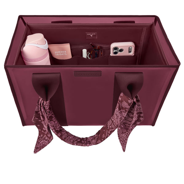 A burgundy tote organizer bag shown from above, filled with travel accessories including a pink tumbler, a phone case, a small box, and a scarf tied to the handles. 
#color_mulberry