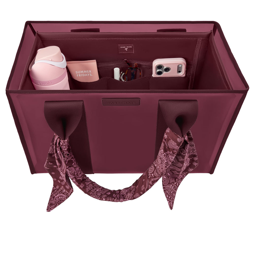 A burgundy tote organizer bag shown from above, filled with travel accessories including a pink tumbler, a phone case, a small box, and a scarf tied to the handles. 
#color_mulberry