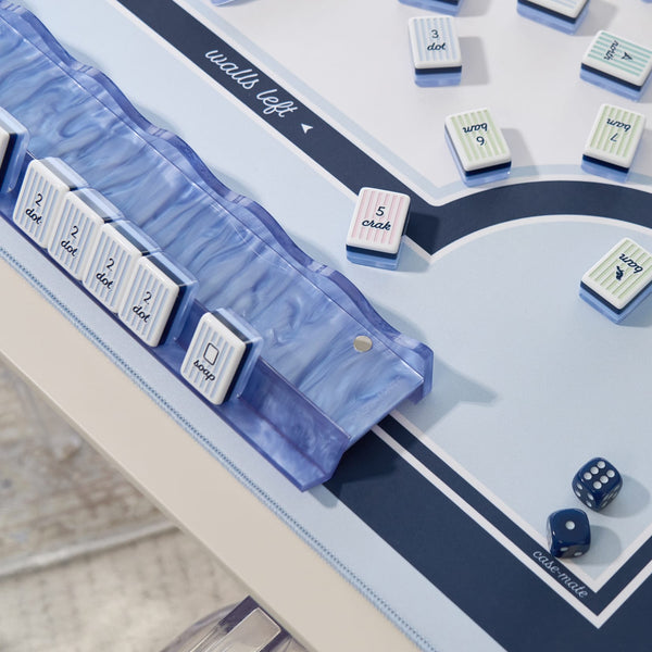 A close-up of a board game setup on a light tabletop, showing a translucent blue marble dice tray, white numbered tiles with blue and pink striped borders, and blue dice.
#color_carolina blue pearl