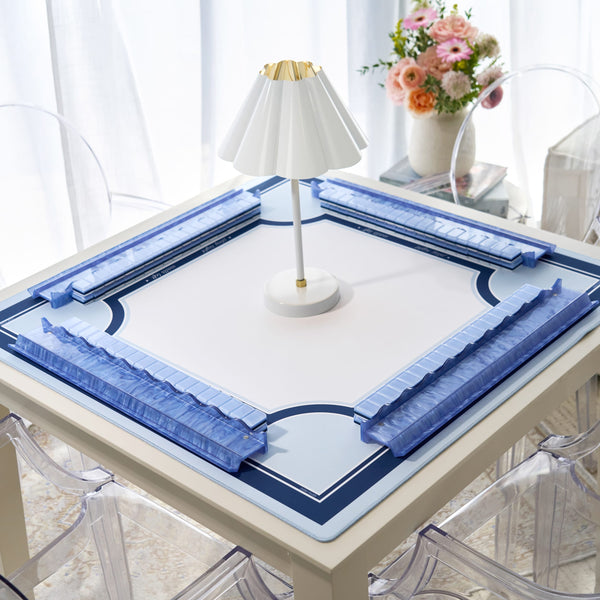 A square white table mat with blue decorative corner sections and a central blue border, displayed on a table in a bright room with a small white lamp and a vase of flowers.
#color_classic carolina blue
