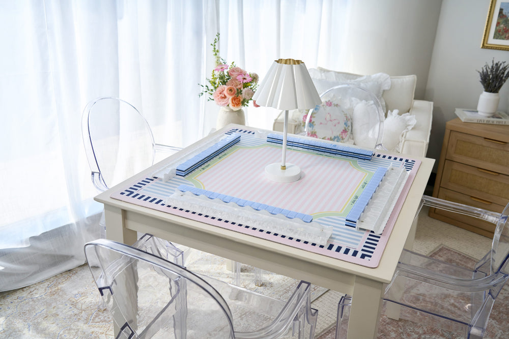A square table covered with a pastel pink and blue patterned mat, surrounded by clear acrylic dining chairs in a bright, softly lit room with white curtains and a lamp.
#color_classic cabana stripe