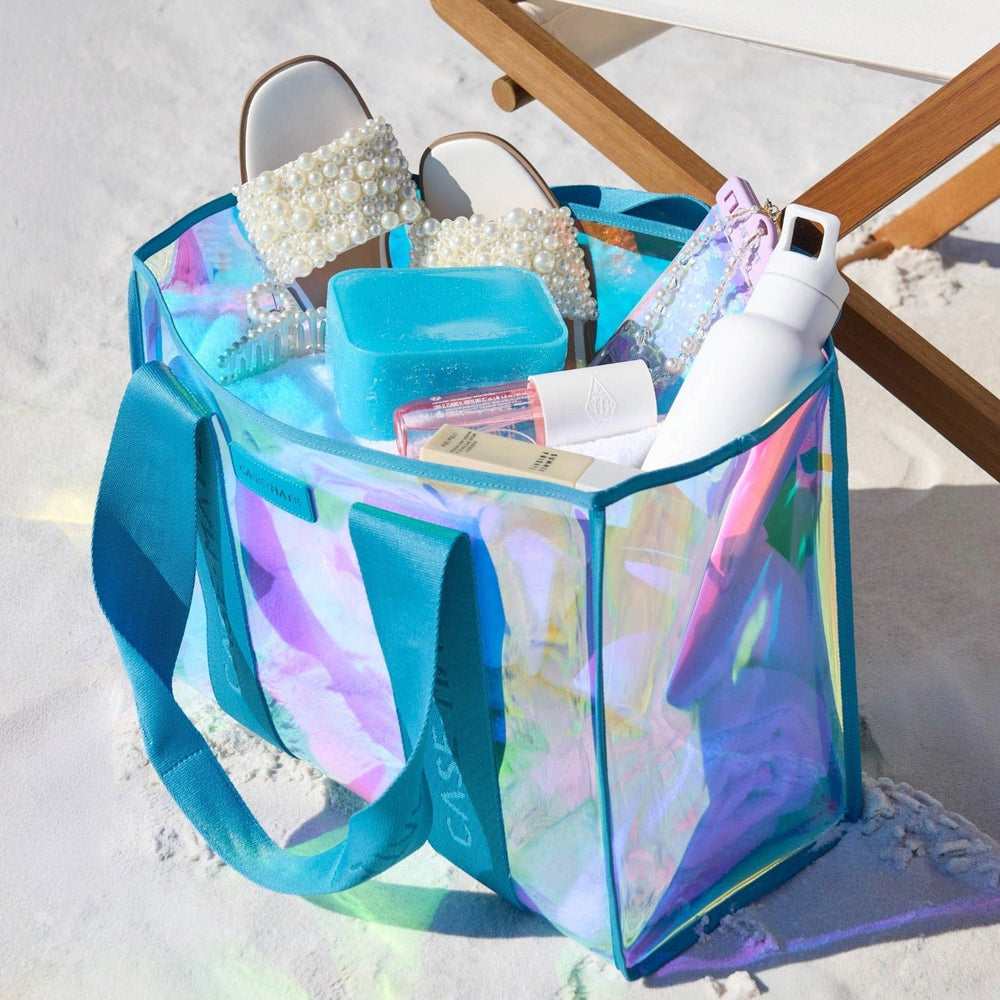 Iridescent beach bag filled with beach accessories on a sandy background 
#color_emerald blue soap bubble®