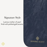 A close-up of a Kendra Scott product with a dark navy blue marbled finish and polished gold accents, including the brand name and logo. Text on the left reads 'Signature Style' and describes the lustrous mother-of-pearl finish with gold accents.