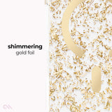 A close-up of a clear or white surface decorated with shimmering gold foil flakes and metallic gold cutout shapes, with the text 'shimmering gold foil' on the left.
#color_karat
