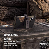 Pelican rugged phone case with integrated black stand, shown holding a smartphone upright on a dark outdoor surface with camouflage-patterned inner panel. Text on image reads 'INTEGRATED STAND PROP IT UP FOR CALLS AND DIRECTIONS WHILE ON THE JOB' and includes Pelican badge.
#color_shield elements hunter camo
