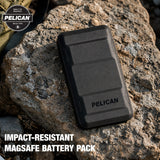A black Pelican impact-resistant MagSafe battery pack resting on a large rock outdoors, with the Pelican logo printed on the front and promotional text at the bottom.