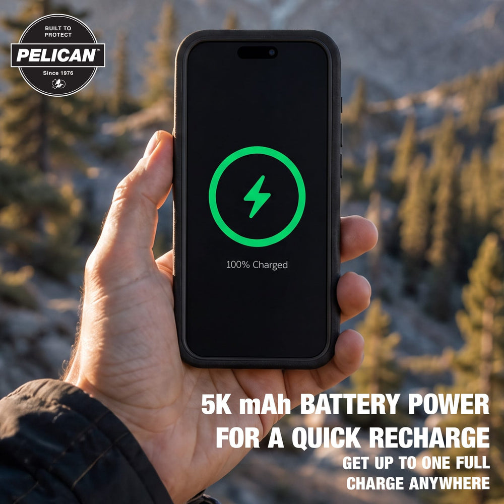 A hand holds a black Pelican smartphone battery charger or power bank in front of a blurred outdoor mountain and forest background. The screen shows a green charging icon and '100% Charged' text.