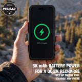 A hand holds a black Pelican smartphone battery charger or power bank in front of a blurred outdoor mountain and forest background. The screen shows a green charging icon and '100% Charged' text.