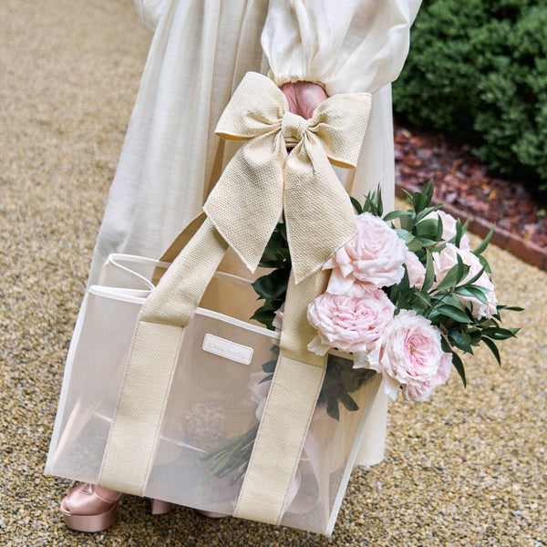 Person holding a gift bag with a large bow and pink flowers, standing on a gravel path.
#color_raffia bow
