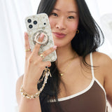A woman holding a smartphone in front of her face, showing a decorative phone case with a built-in ring stand and a pearl charm strap. The case has a white background with a gold abstract line pattern.
#color_floral gems