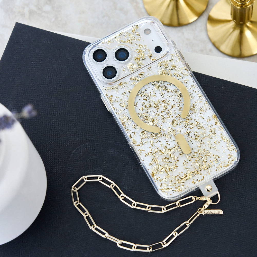 A clear phone case with gold foil flakes and a large gold circular MagSafe ring, shown on a phone with a matching gold chain strap, styled on a dark surface with gold candle bases in the background.

#color_karat

