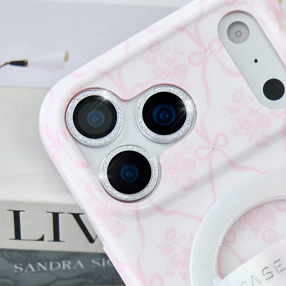 A close-up of a white phone case with a light pink floral pattern and silver rhinestone-trimmed camera rings, shown resting on a light-colored book.

#color_twinkle
