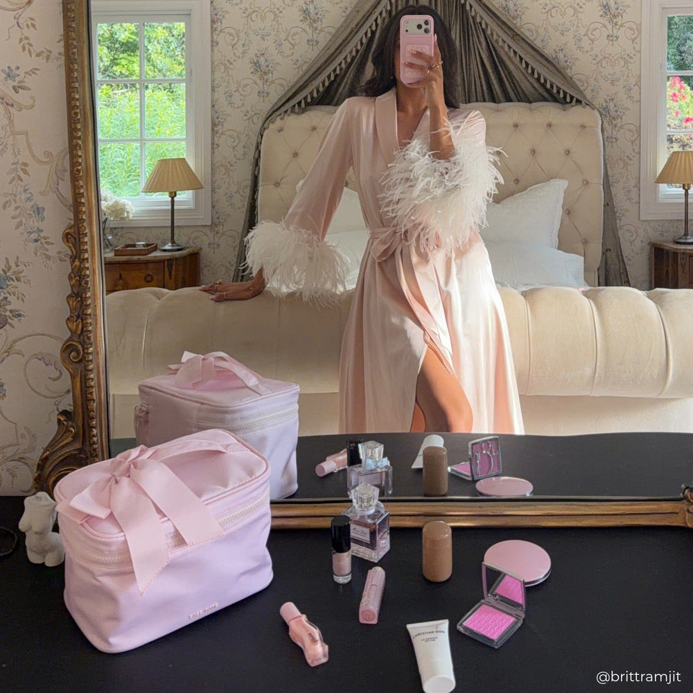 Person in a robe taking a mirror selfie with makeup and a pink bag on a table.