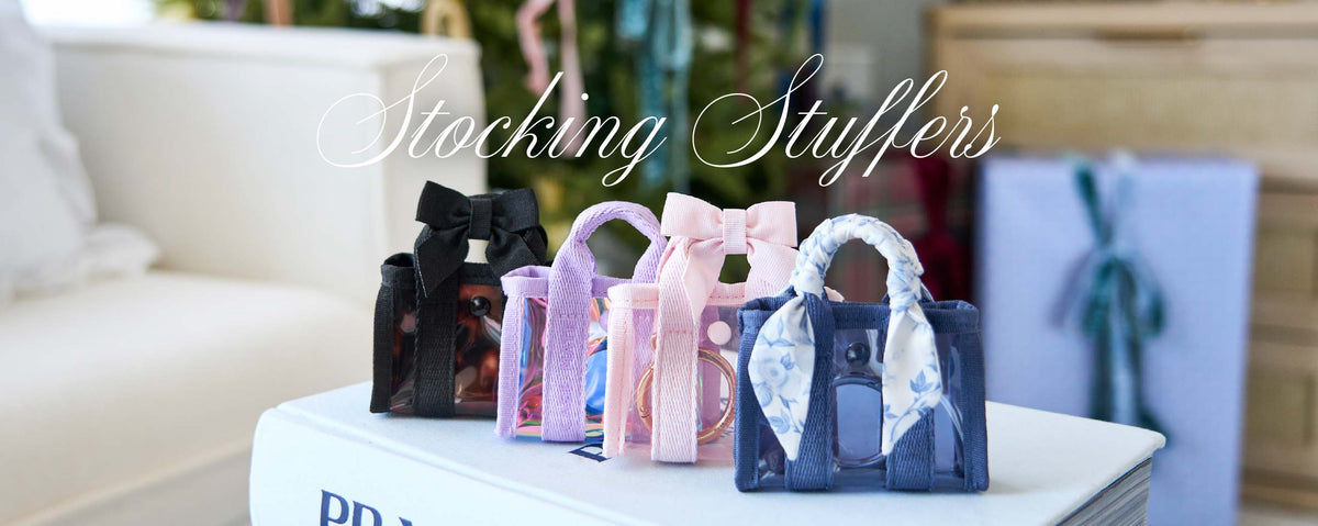 Three small transparent handbag-shaped stocking stuffers in black, pink, and navy blue are lined up on a white book, with a white sofa and a gift box blurred in the background. The text 'Stocking Stuffers' is written across the top.