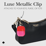Kate Spade metallic clip accessory shown attached to a black leather bag, with text reading 'Luxe Metallic Clip' and 'ATTACHES TO YOUR KEYS, PURSE, OR TOTE'.