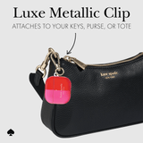 Kate Spade black textured leather bag with a gold-tone metallic clip holding a pink and red round keychain, shown on a light gray background with text reading 'Luxe Metallic Clip' and 'ATTACHES TO YOUR KEYS, PURSE, OR TOTE.'.