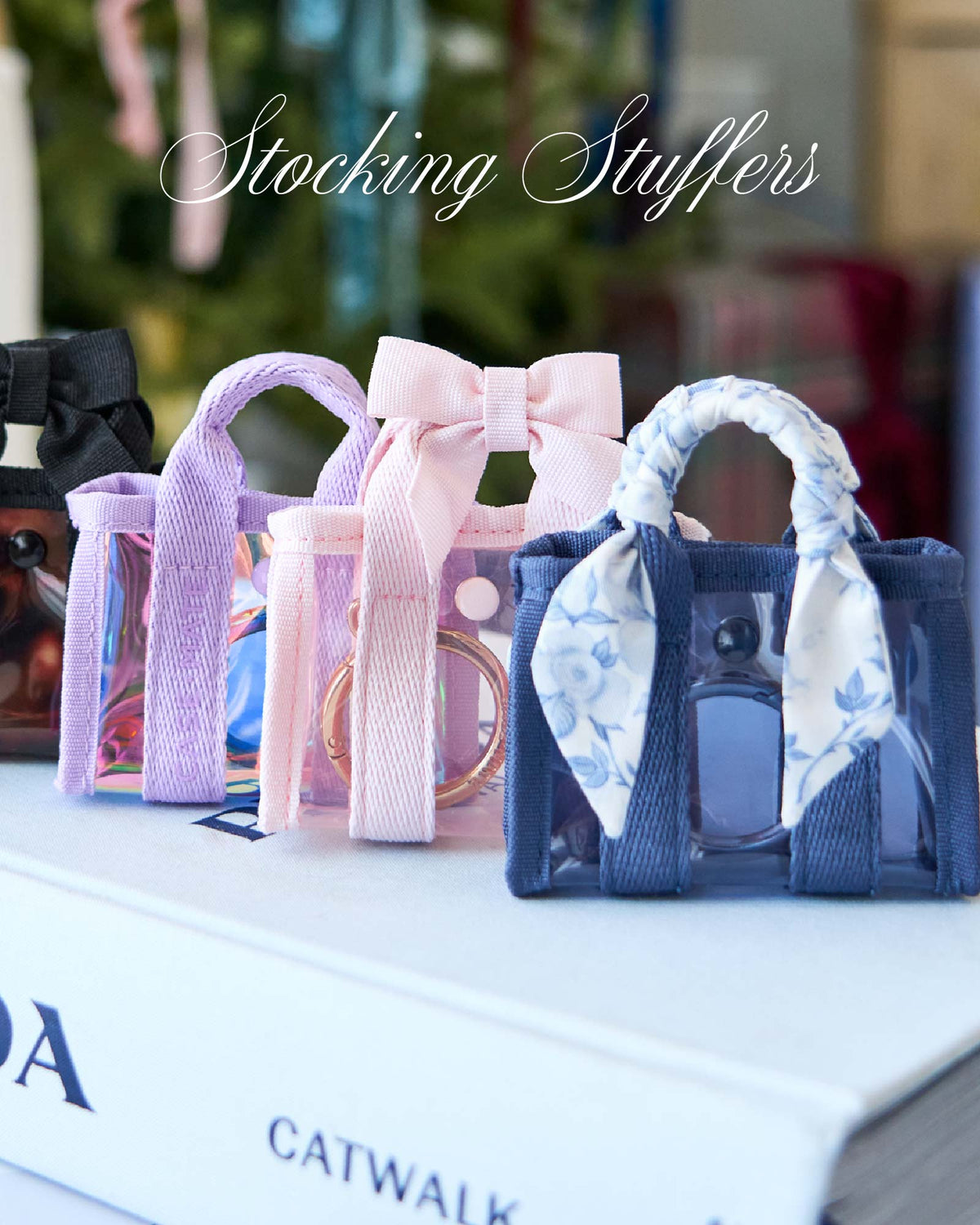 Three small transparent handbag-shaped stocking stuffers in black, pink, and navy blue are lined up on a white book, with a white sofa and a gift box blurred in the background. The text 'Stocking Stuffers' is written across the top.