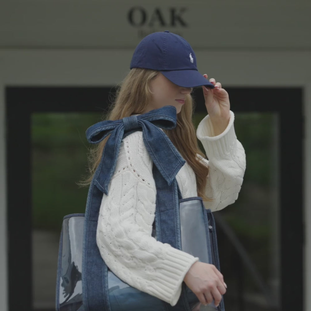 Woman in an outdoor setting with a blue baseball hat and denim strap jelly tote bag on her shoulder #color_dark wash denim bow