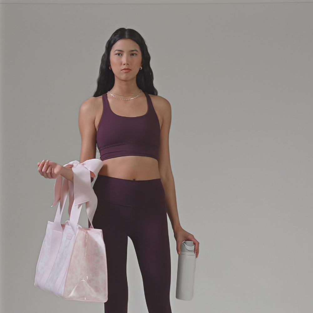 A woman in a plum workout outfit showing off her jelly tote bag and how big it is with. a water bottle
#color_ballet pink afternoon rosé bow