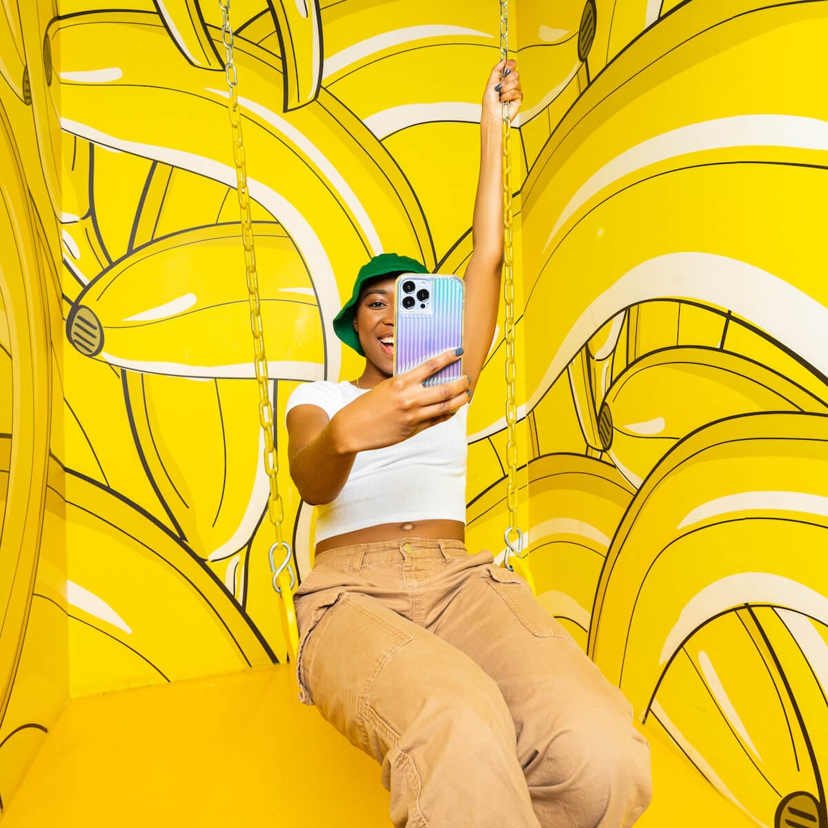 Woman swinging on a swing in a banana room taking a selfie with Tough Groove. color::Iridescent