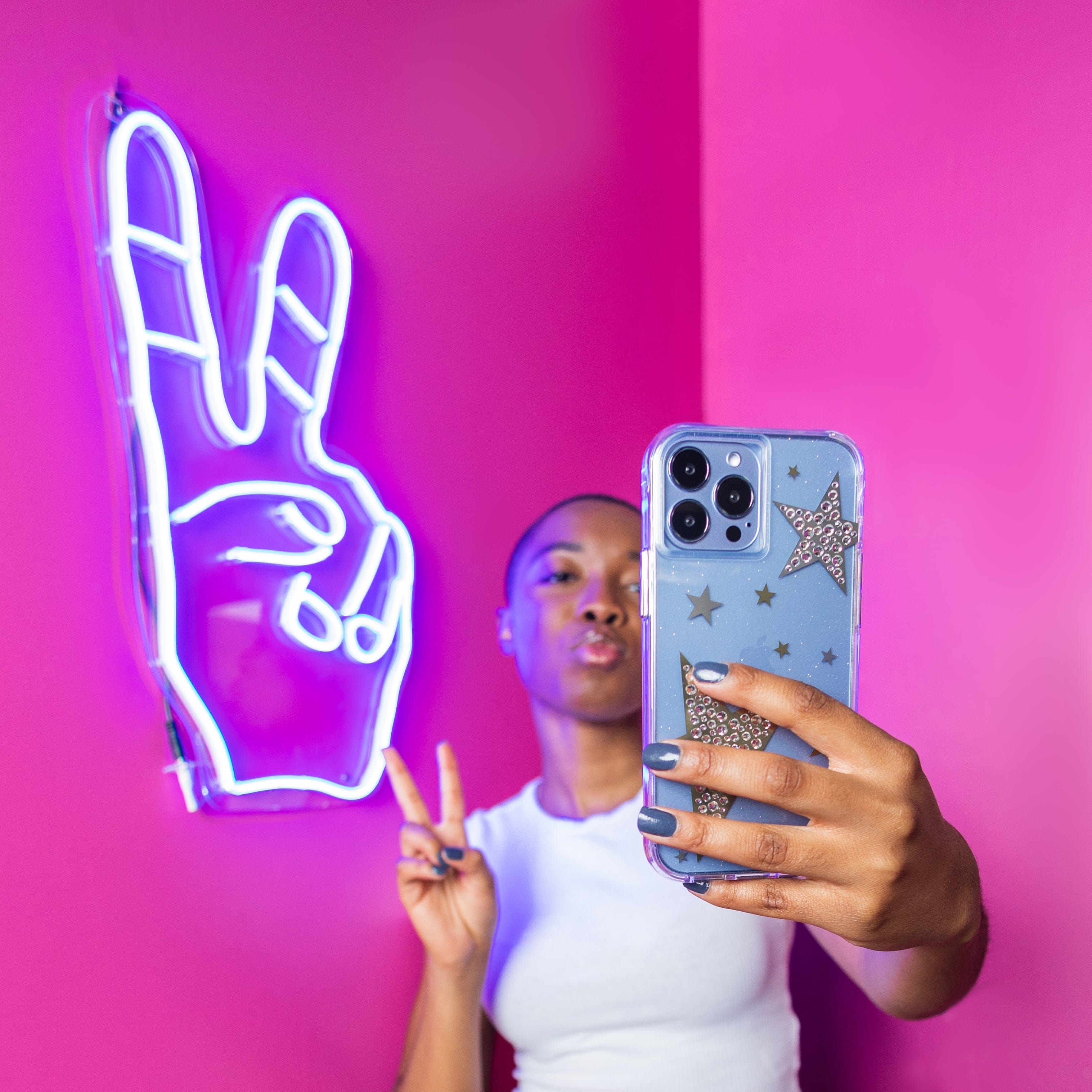 Woman taking selfie in front of peace sign neon light holding a phone with Sheer Superstar. color::Sheer Superstar