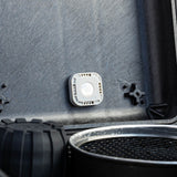 White square AirTag sticker mount with adhesive backing mounted on grey vehicle surface.