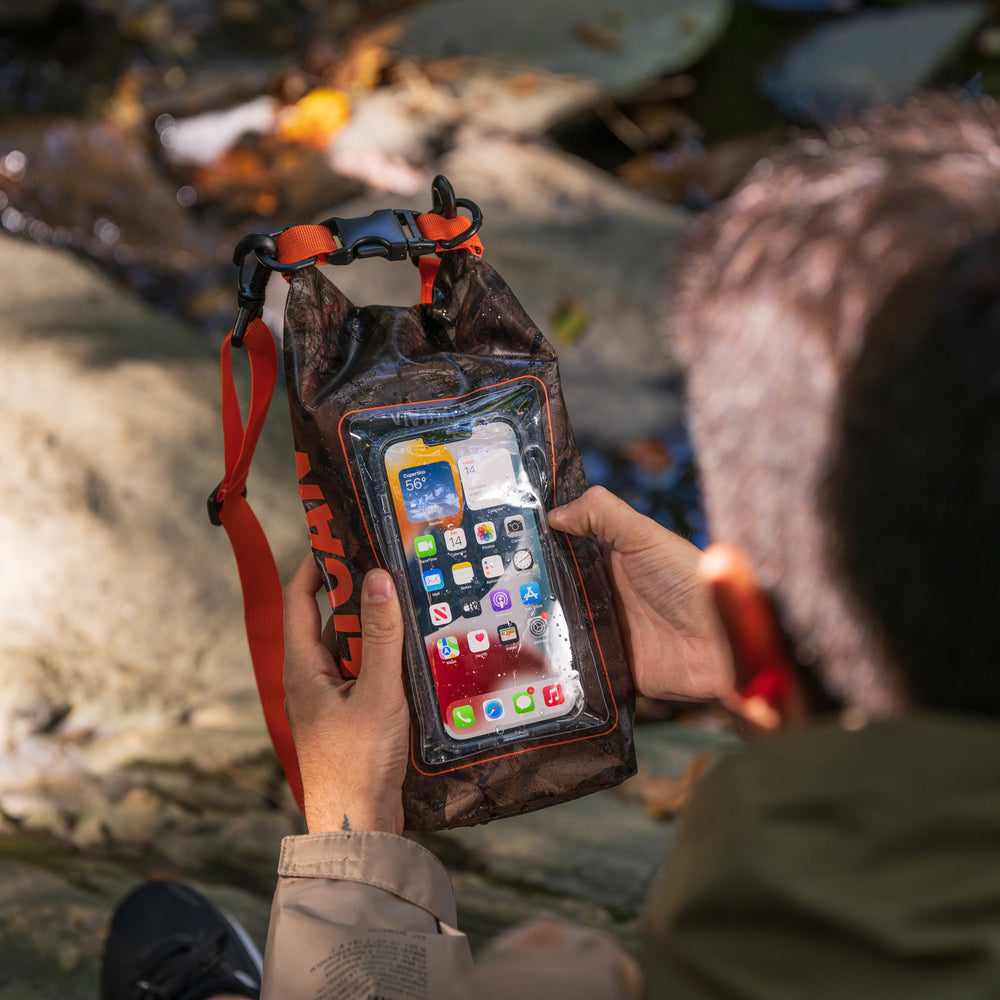 Hunter camo dry bag with red shoulder strap held in hand displaying smartphone inside clear window, near water.